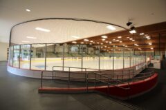 Paul Hruby Ice Arena features a clean ice rink with spectator seating and a protective net overhead. A ramp provides access.